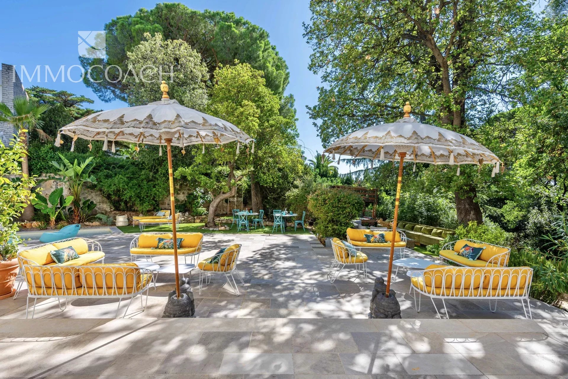 Sunny outdoor lounge area with yellow-cushioned white metal sofas under decorative umbrellas on a stone patio, surrounded by green trees and plants.