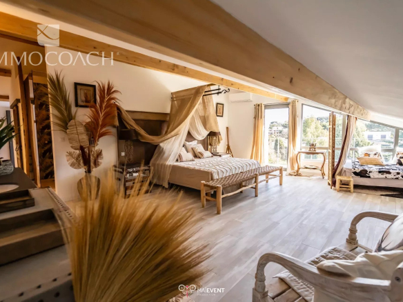 Luxurious loft bedroom with a four-poster bed under a sheer canopy, a woven bench at the foot, and sunlit windows opening to a balcony.