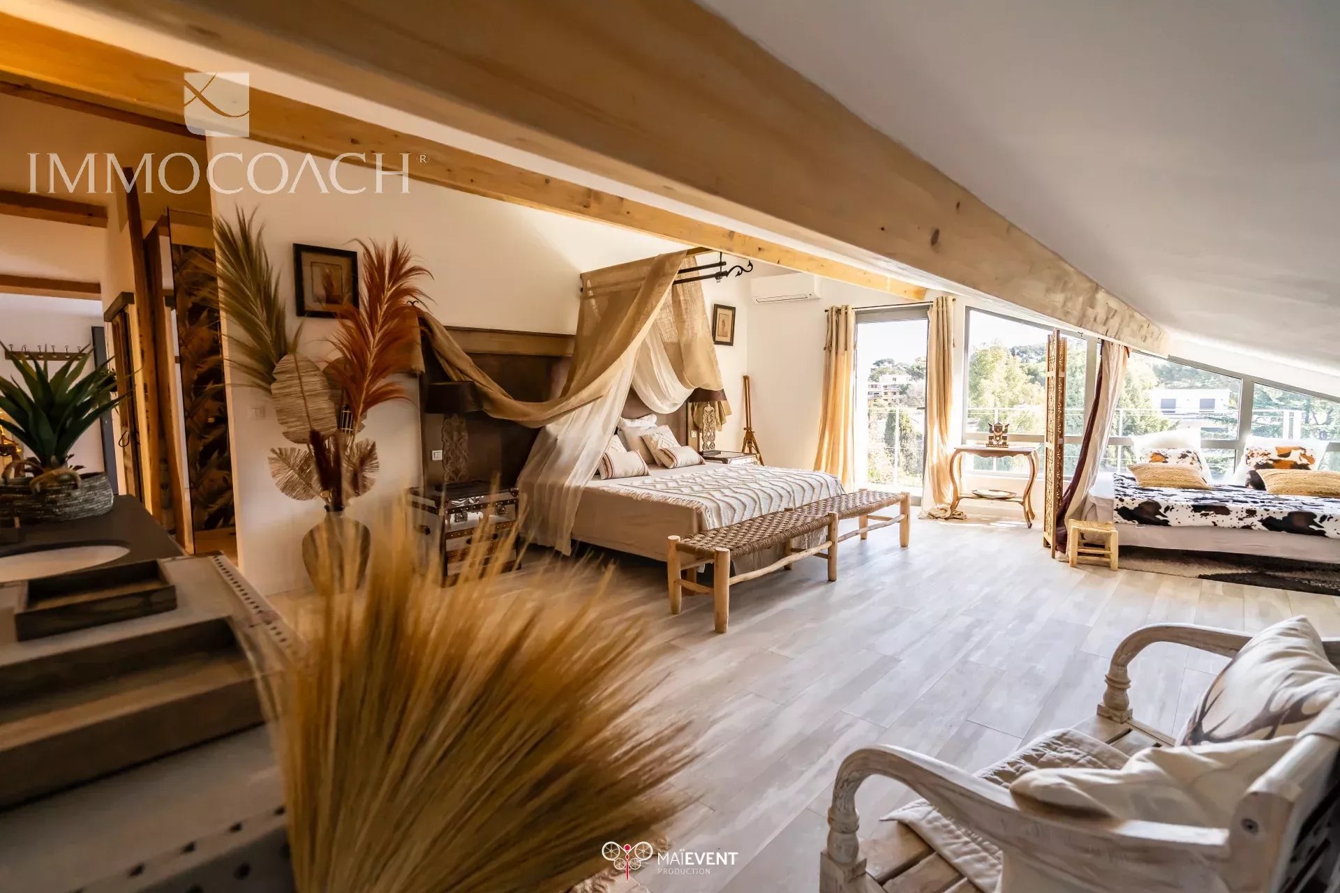 Luxurious loft bedroom with a four-poster bed under a sheer canopy, a woven bench at the foot, and sunlit windows opening to a balcony.