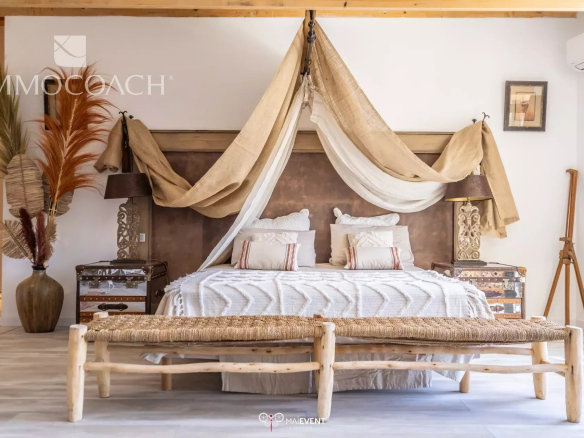 Boho-inspired bedroom with a canopied bed, beige drapes, and white bedding flanked by ornate lamps and vintage trunks.