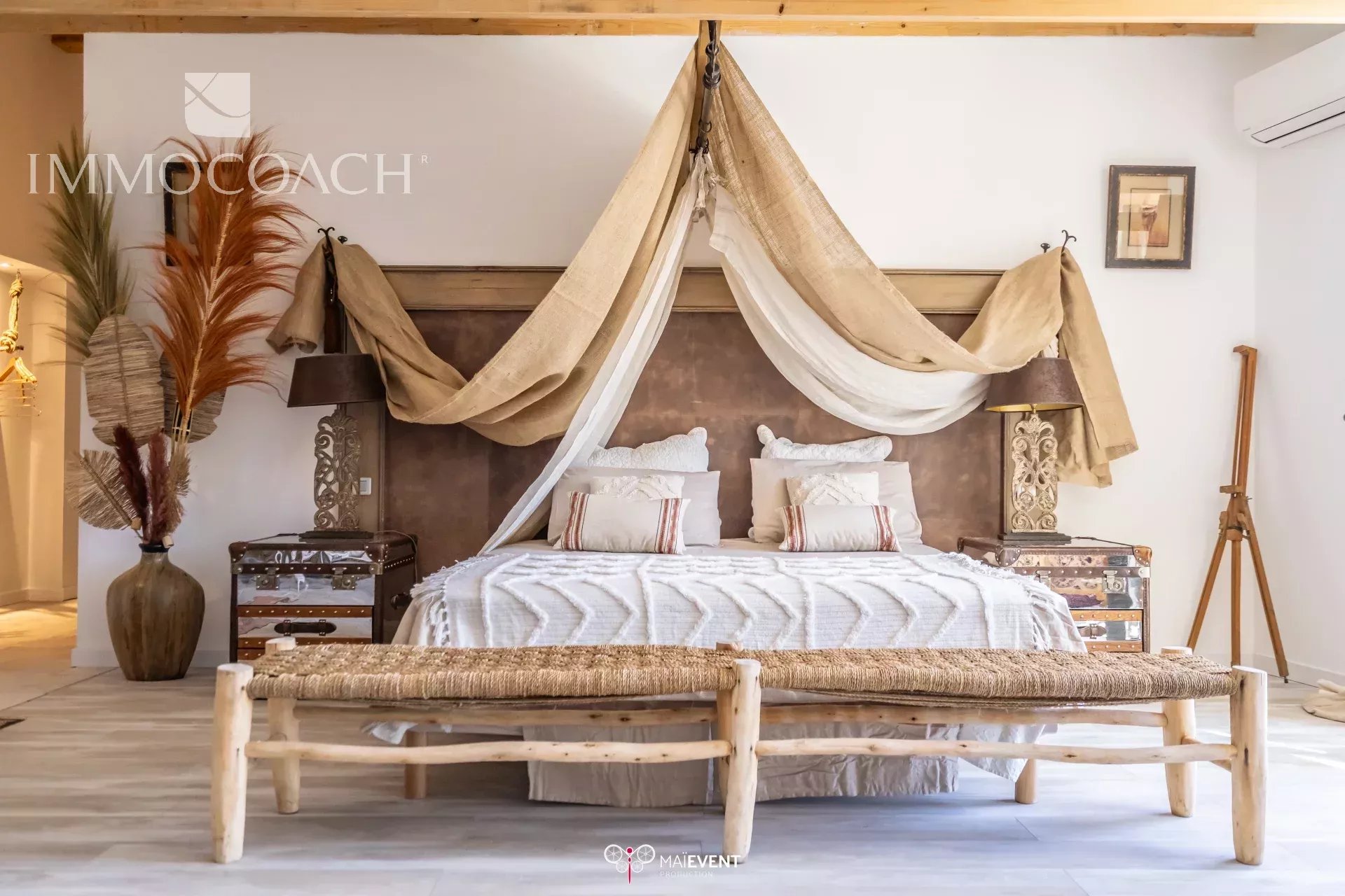 Boho-inspired bedroom with a canopied bed, beige drapes, and white bedding flanked by ornate lamps and vintage trunks.