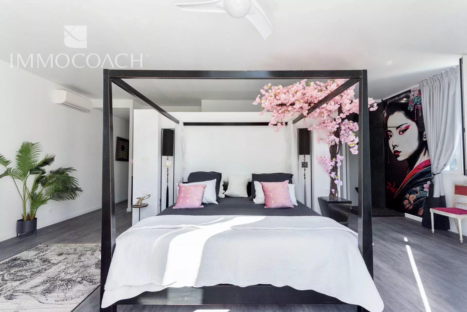 Modern bedroom with a black four-poster bed, white bedding, and pink accent pillows under a bright, airy showroom glow.