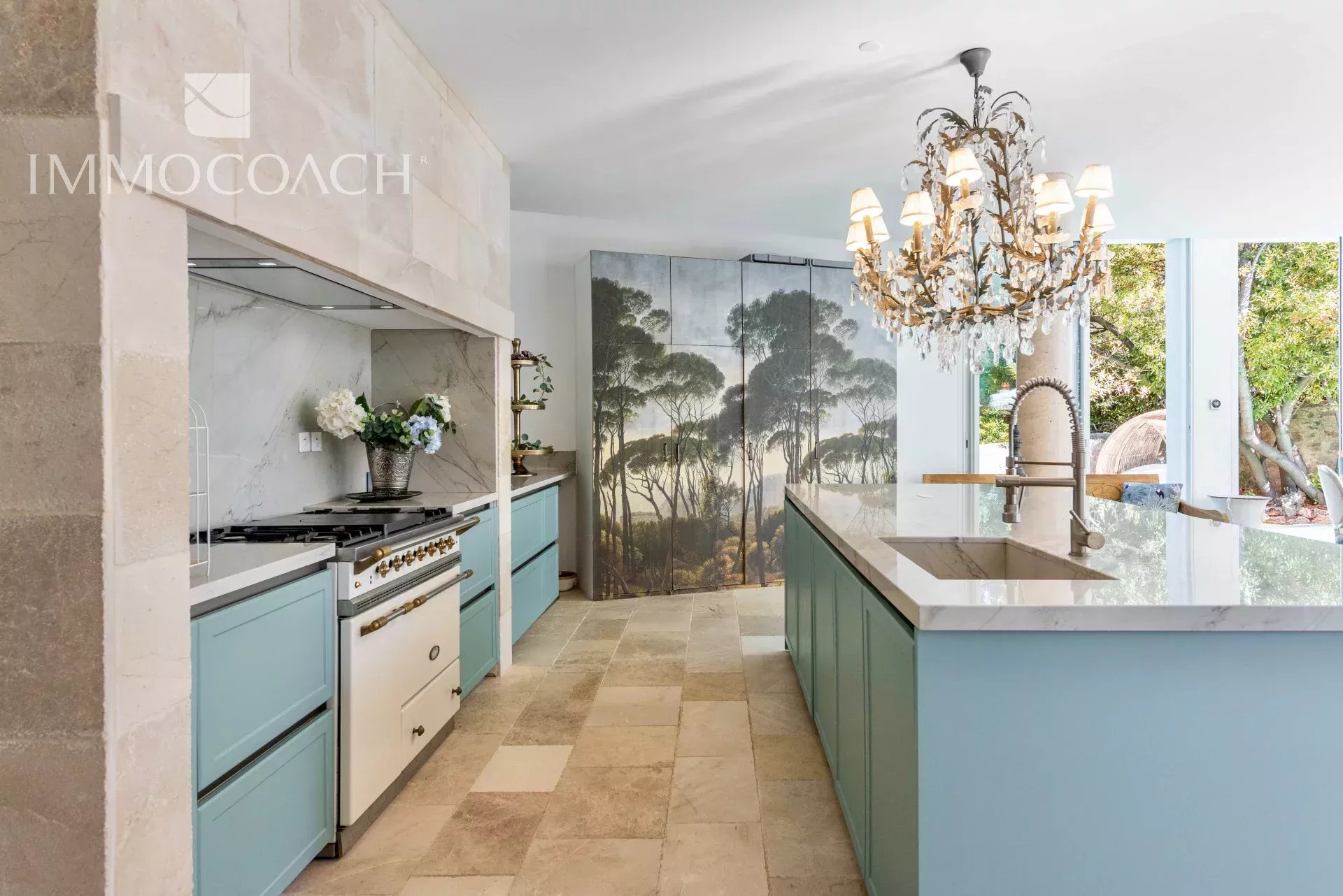 Bright kitchen with blue cabinets, marble countertops, and a large island featuring a modern brass faucet and crystal chandelier.