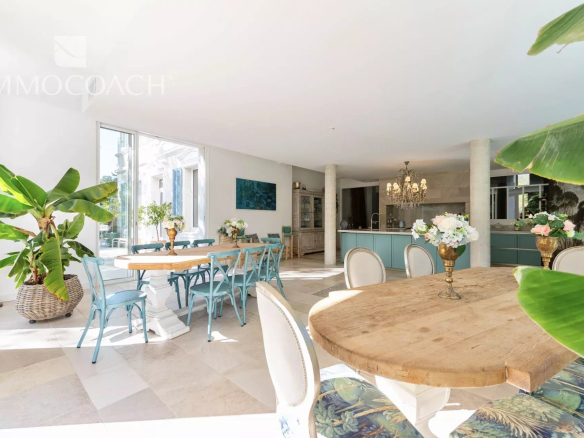 Bright open-plan dining area with a round wooden table, blue chairs, a turquoise kitchen island, chandelier, and large green plants.