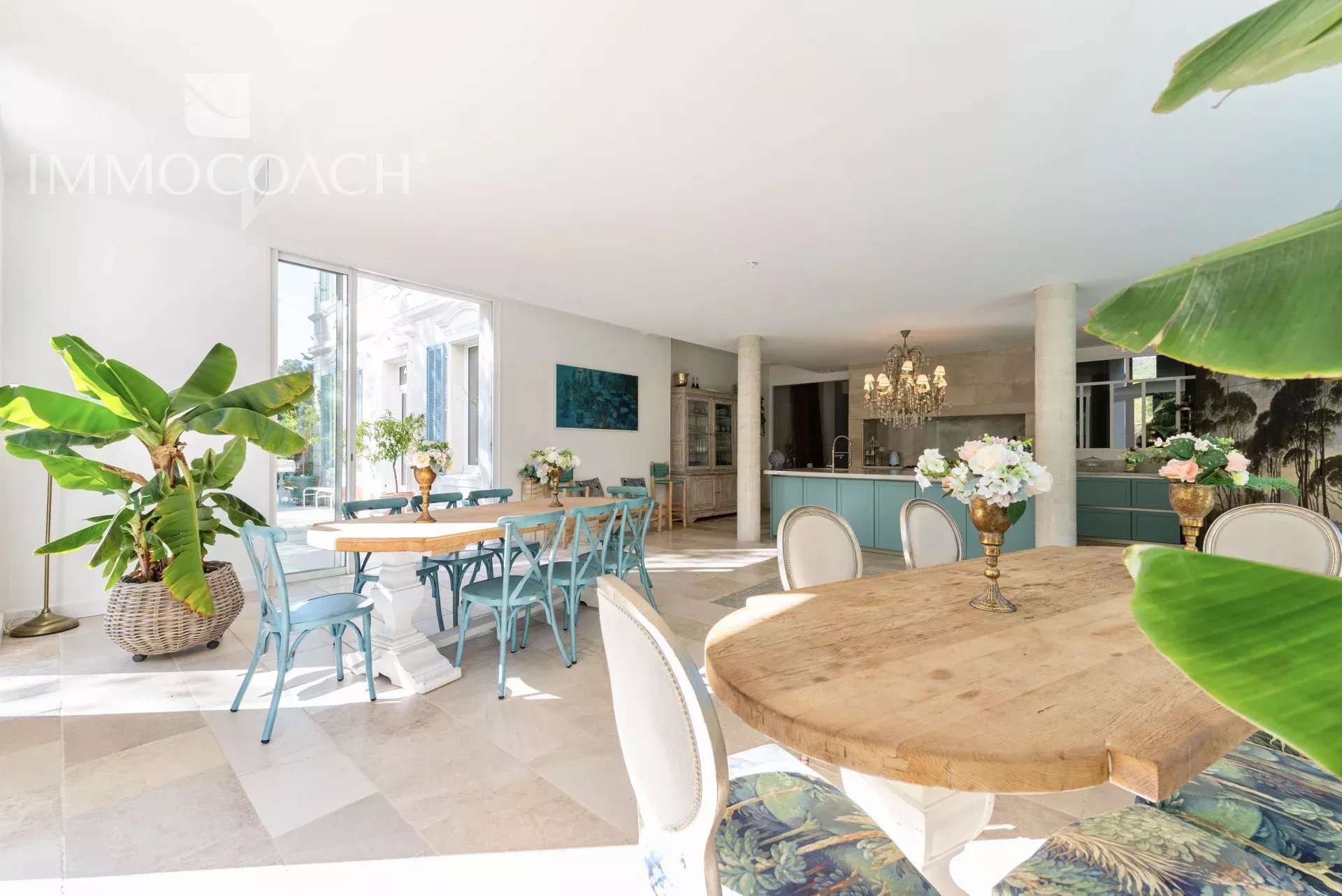 Bright open-plan dining area with a round wooden table, blue chairs, a turquoise kitchen island, chandelier, and large green plants.