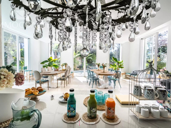 Bright dining room with a large crystal chandelier, round tables, pastel chairs, and tall windows with greenery.