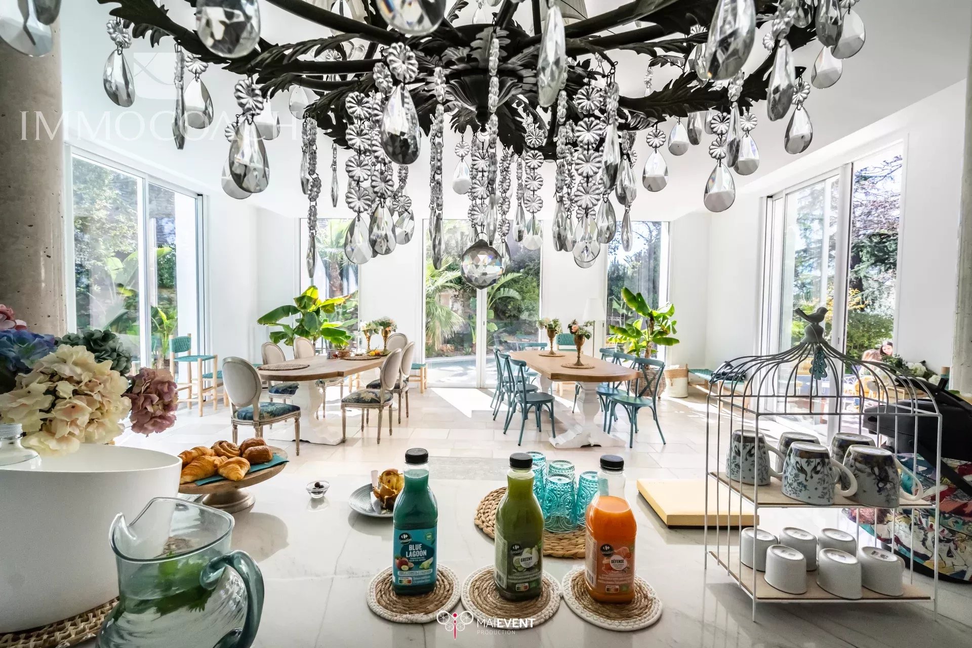 Bright dining room with a large crystal chandelier, round tables, pastel chairs, and tall windows with greenery.