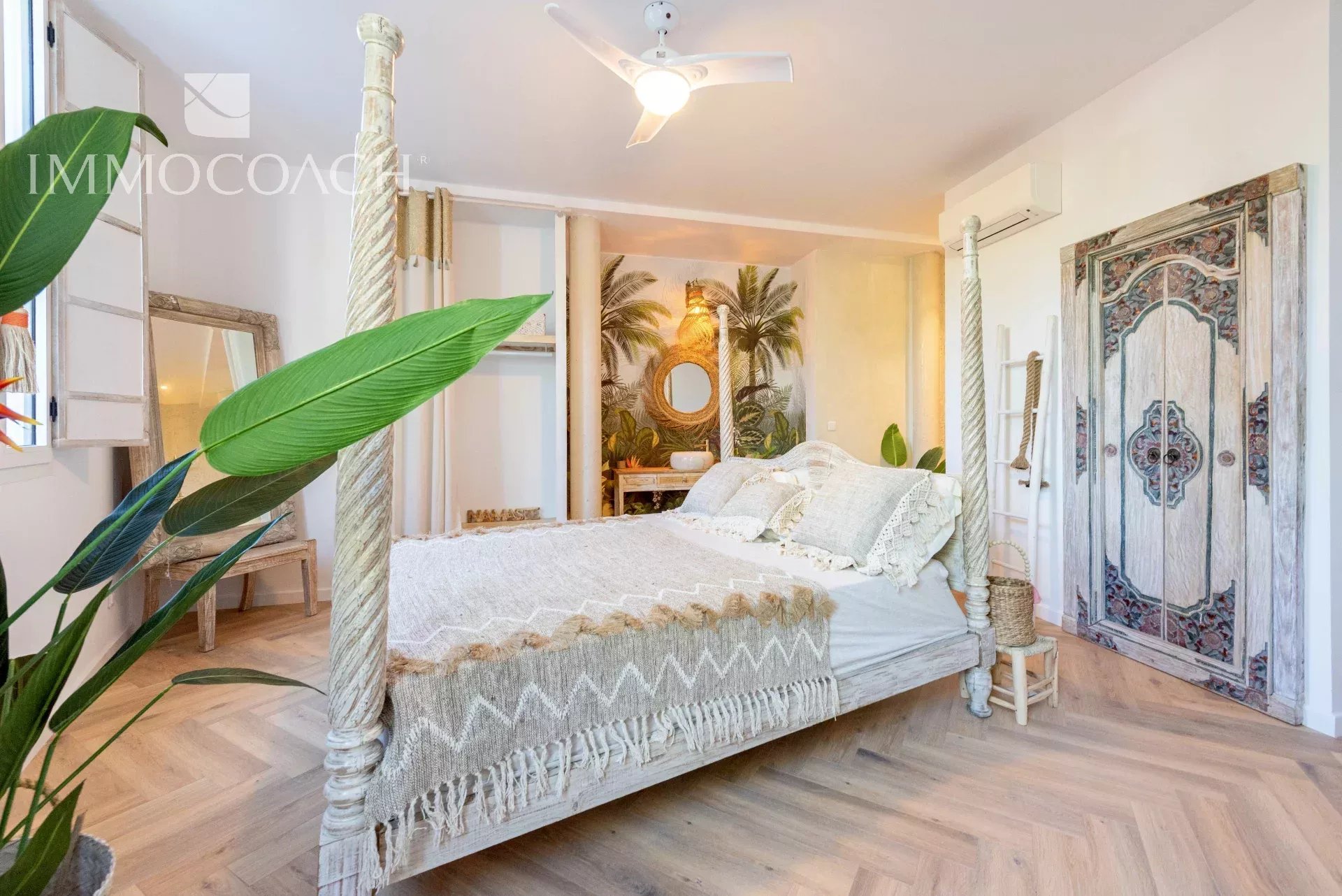 Boho-style four-poster bed with a cream knitted throw in a bright tropical-themed bedroom with plants and wicker accents.