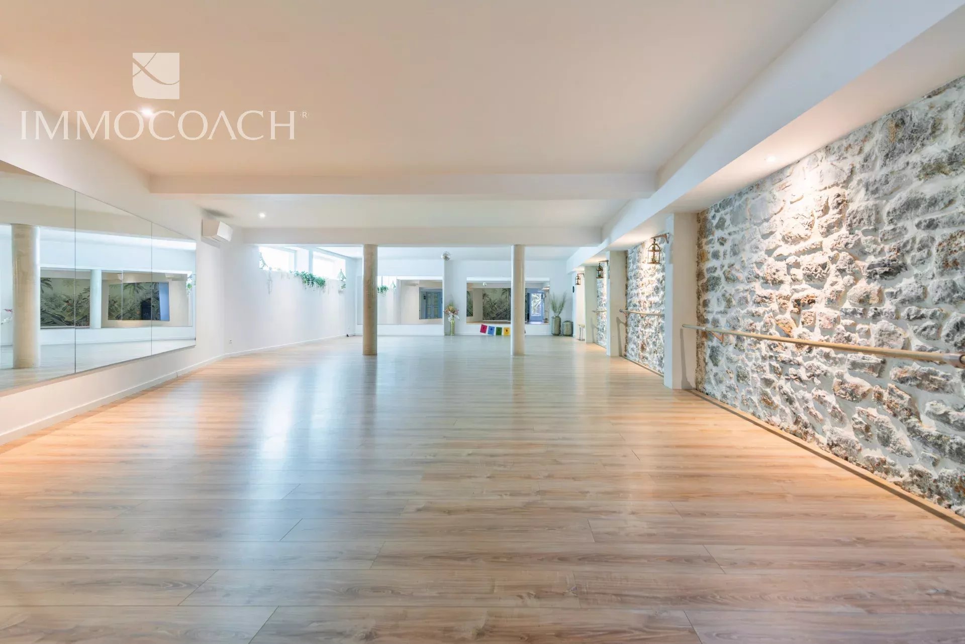 Spacious dance or fitness studio with a mirrored wall on the left, stone wall with a barre on the right, and warm wooden flooring.