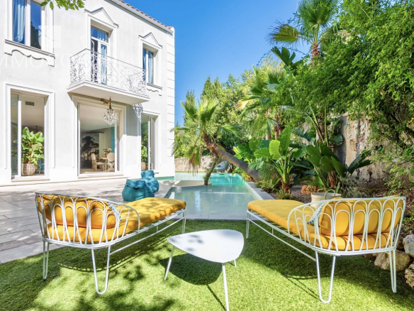 White villa with ornate balcony and glass doors, outdoor seating by a small pool amid tropical plants.