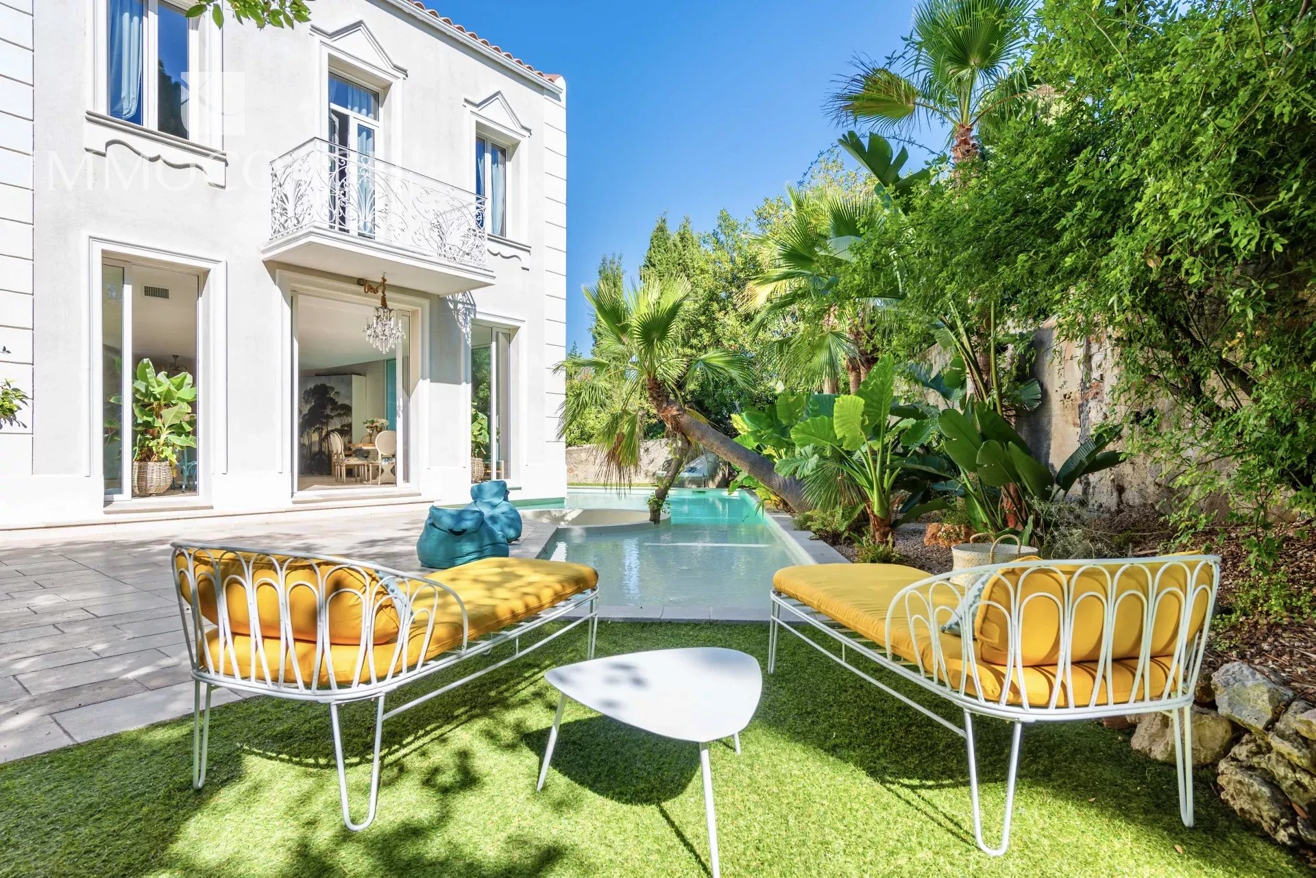 White villa with ornate balcony and glass doors, outdoor seating by a small pool amid tropical plants.
