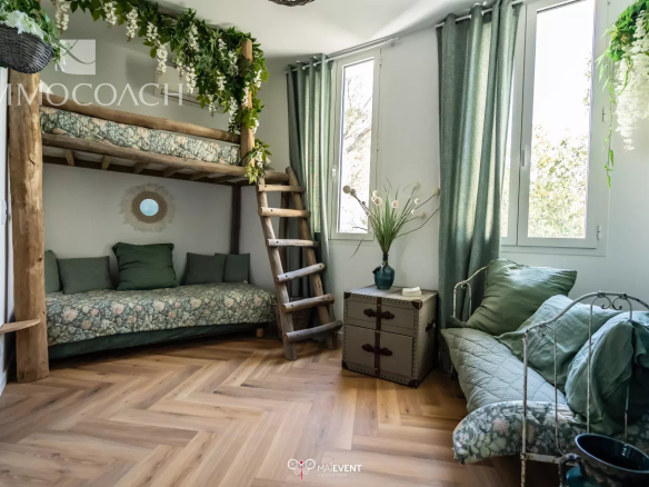 Rustic living space with a wooden bunk bed, green floral bedding, and a ladder, plus a green-cushioned sofa by large windows. Plants hang from the ceiling for a lush, cozy look.