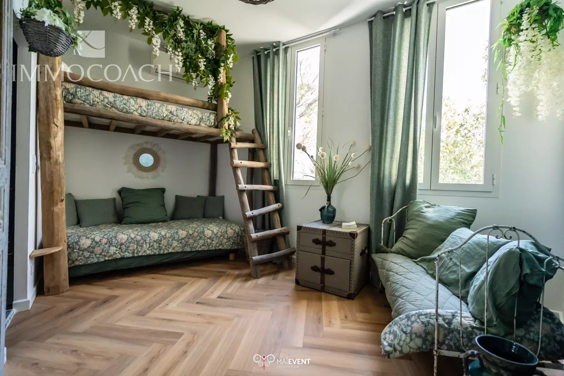 Rustic living space with a wooden bunk bed, green floral bedding, and a ladder, plus a green-cushioned sofa by large windows. Plants hang from the ceiling for a lush, cozy look.