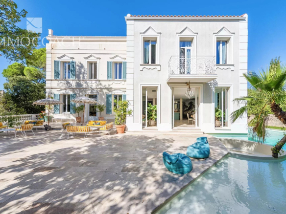White two-story villa with decorative windows, courtyard seating with yellow cushions, and a turquoise pool under a blue sky.