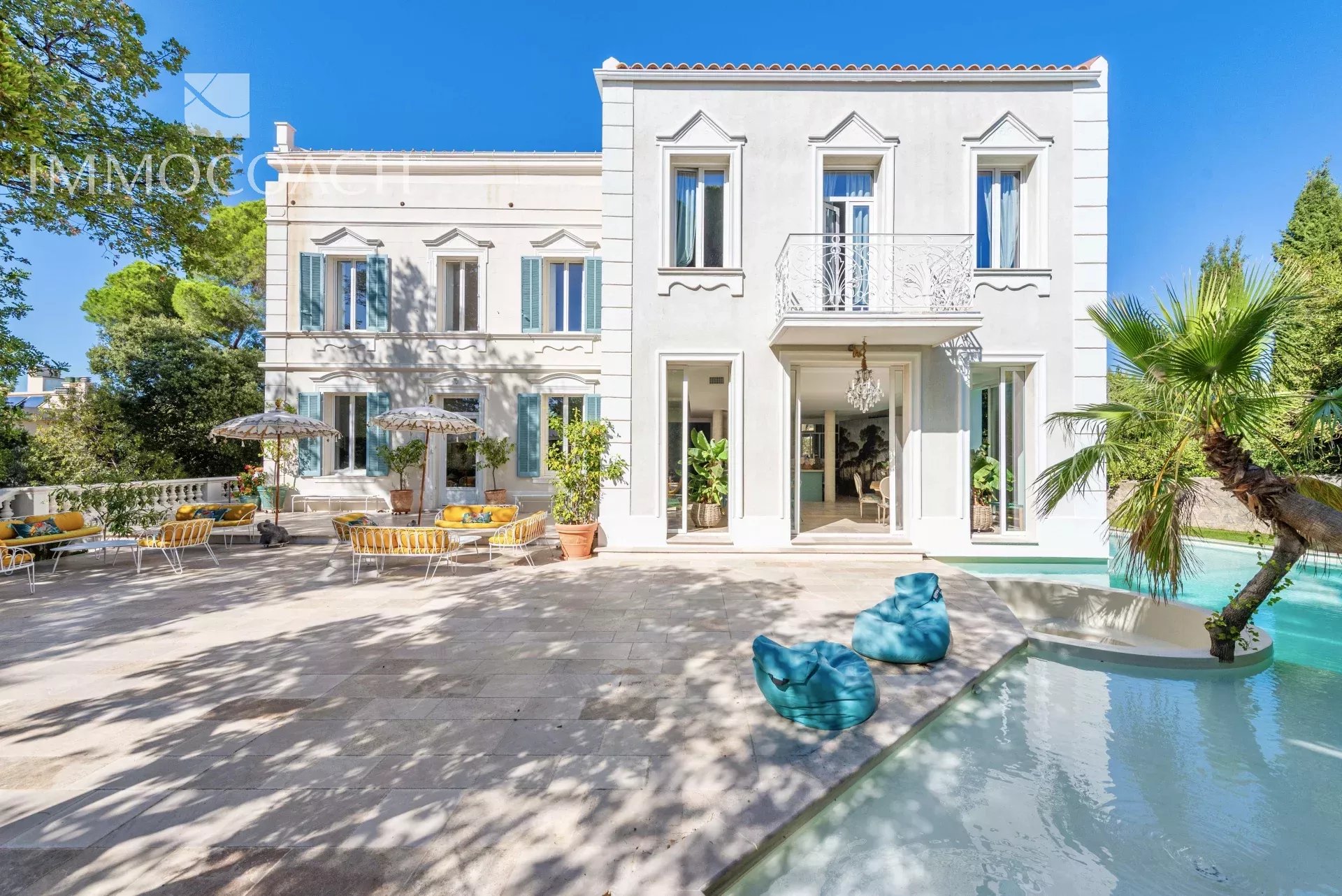 White two-story villa with decorative windows, courtyard seating with yellow cushions, and a turquoise pool under a blue sky.