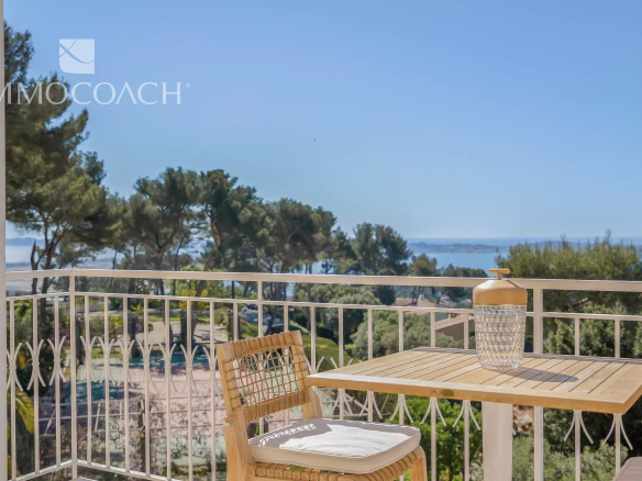 Balcony scene with a wooden table and chair, a decorative glass jar on the table, and a white railing with a sea view in the distance.