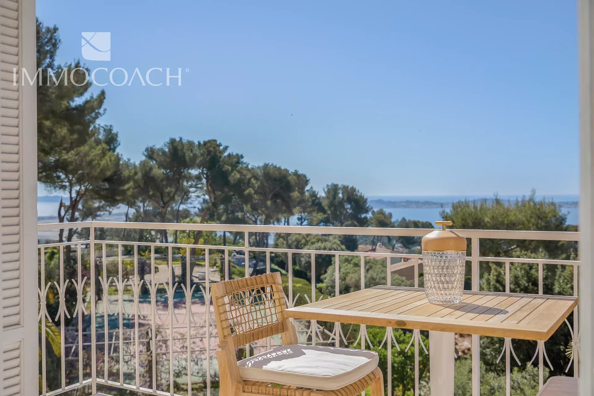 Balcony scene with a wooden table and chair, a decorative glass jar on the table, and a white railing with a sea view in the distance.