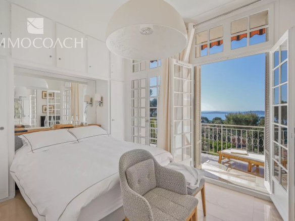 Bright white bedroom opening to a sea-view balcony through tall French doors with a blue horizon beyond the railing.