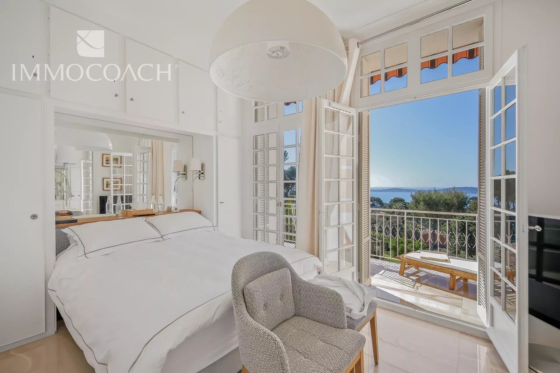 Bright white bedroom opening to a sea-view balcony through tall French doors with a blue horizon beyond the railing.