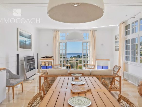 Bright open living and dining area with a long wooden table, wicker chairs, and an ocean view through open French doors nearby.