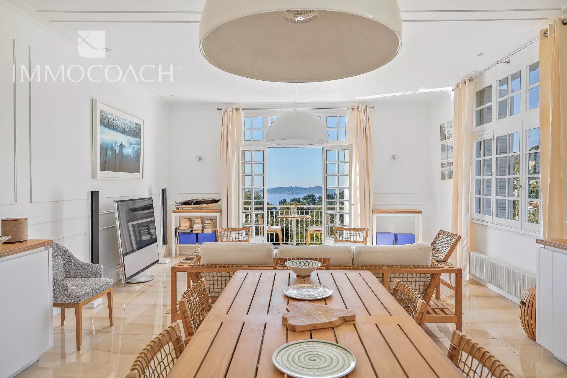 Bright open living and dining area with a long wooden table, wicker chairs, and an ocean view through open French doors nearby.