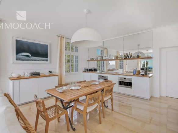 Bright, modern kitchen and dining area with white cabinets, a long wooden table for six, and woven chairs under a large pendant light.