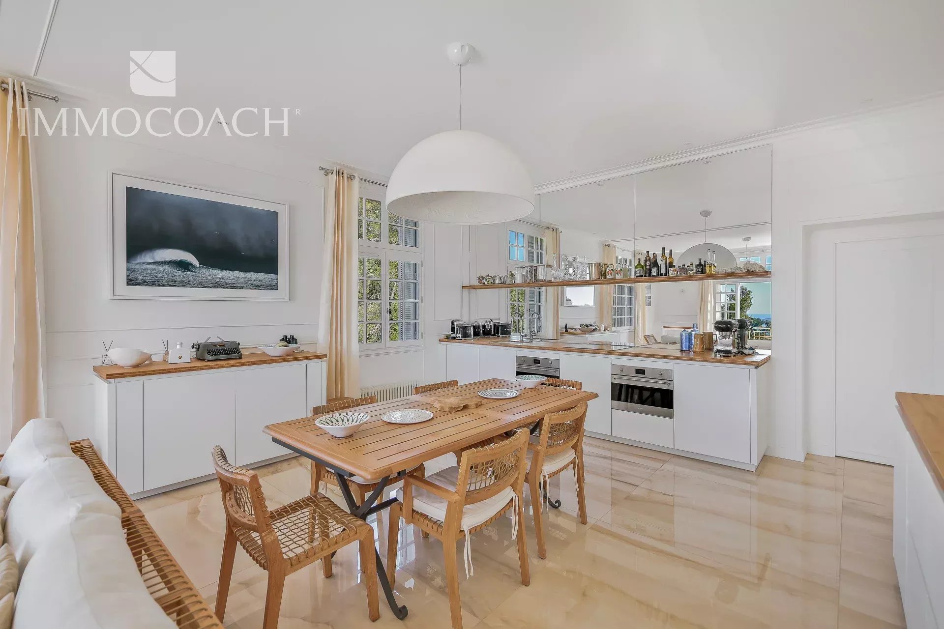 Bright, modern kitchen and dining area with white cabinets, a long wooden table for six, and woven chairs under a large pendant light.