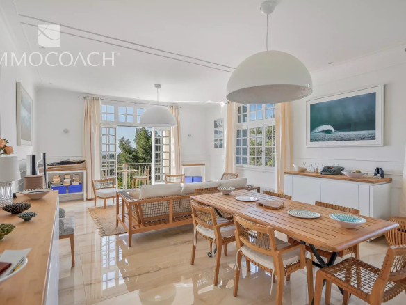 Bright open-plan living and dining room with a long wooden table, woven chairs, and large windows/French doors bringing in natural light.