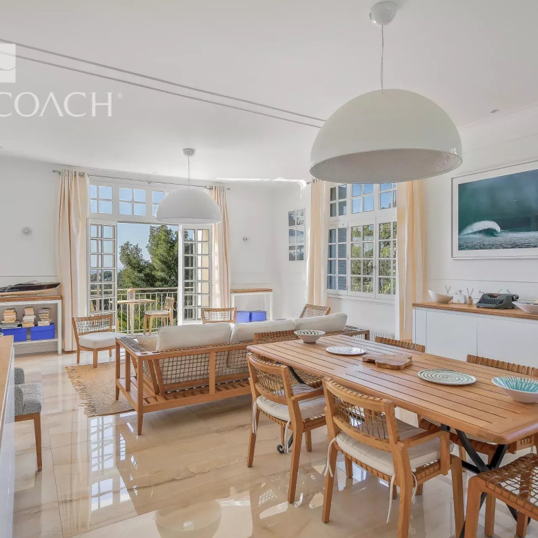 Bright open-plan living and dining room with a long wooden table, woven chairs, and large windows/French doors bringing in natural light.