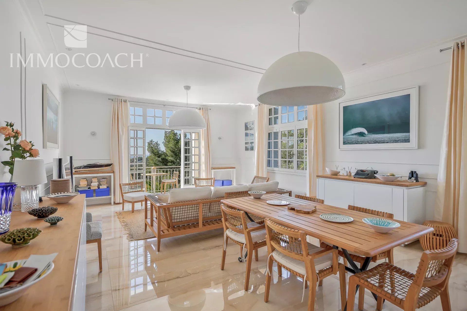 Bright open-plan living and dining room with a long wooden table, woven chairs, and large windows/French doors bringing in natural light.