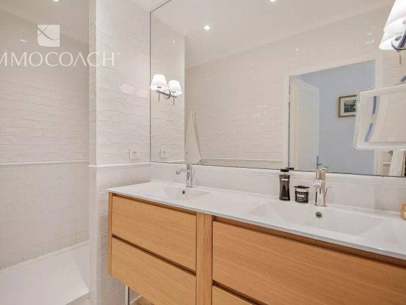 Modern white-tiled bathroom with a double sink vanity, light wood drawers, and a large mirror.