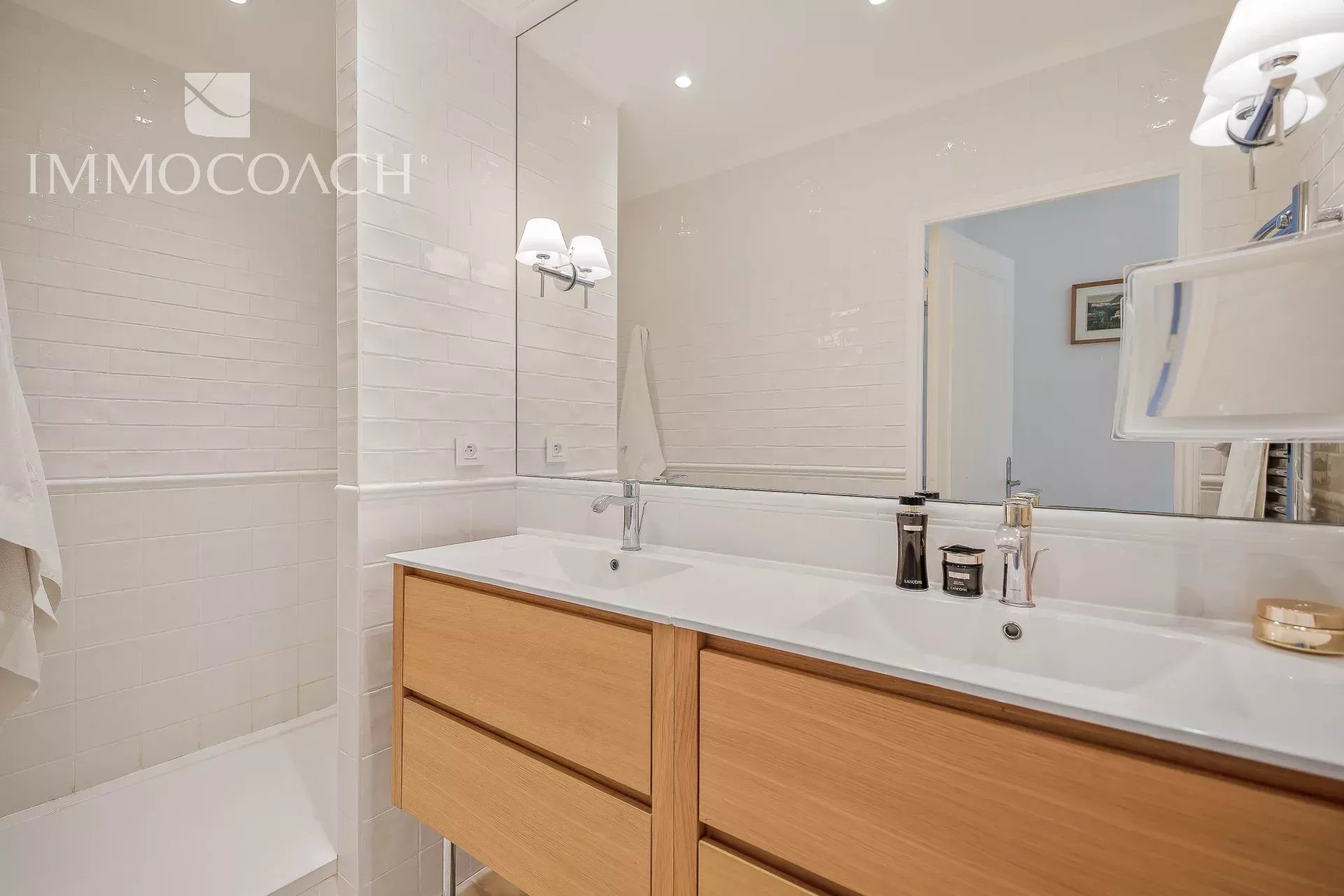 Modern white-tiled bathroom with a double sink vanity, light wood drawers, and a large mirror.