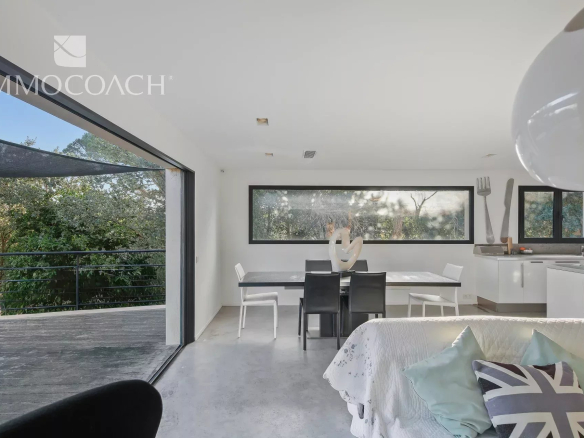 Modern interior of a house with open-plan dining room and kitchen, large windows overlooking the outside.