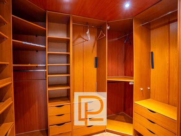 Wooden walk-in closet with open shelves, hanging rods, and multiple drawers along the walls, well-lit by a ceiling light.
