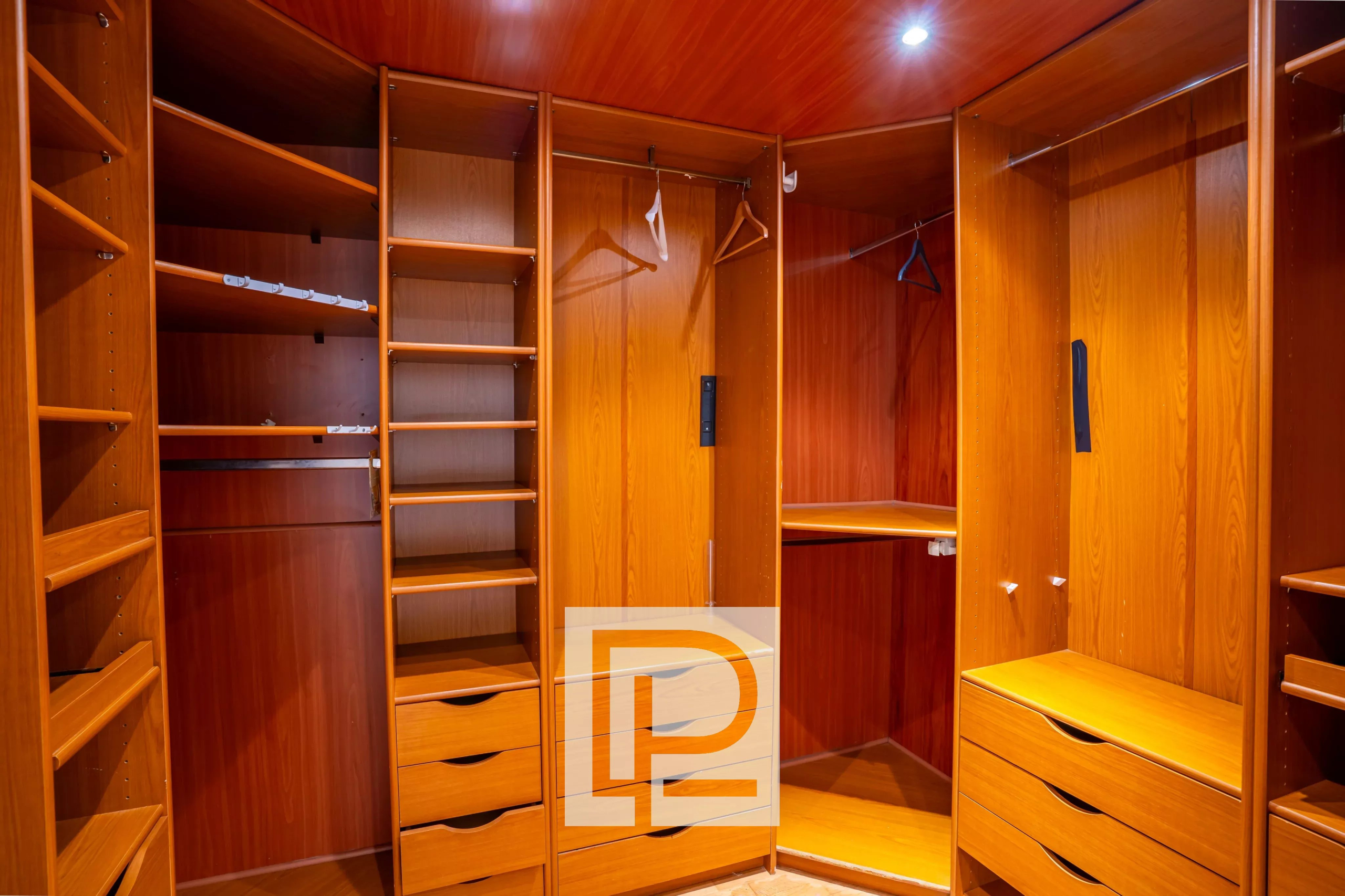 Wooden walk-in closet with open shelves, hanging rods, and multiple drawers along the walls, well-lit by a ceiling light.