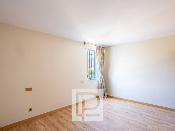 Empty beige-walled room with a light wood floor, a window with bars, and a patterned curtain pulled to the side on the far wall, plus a wooden dresser on the right.