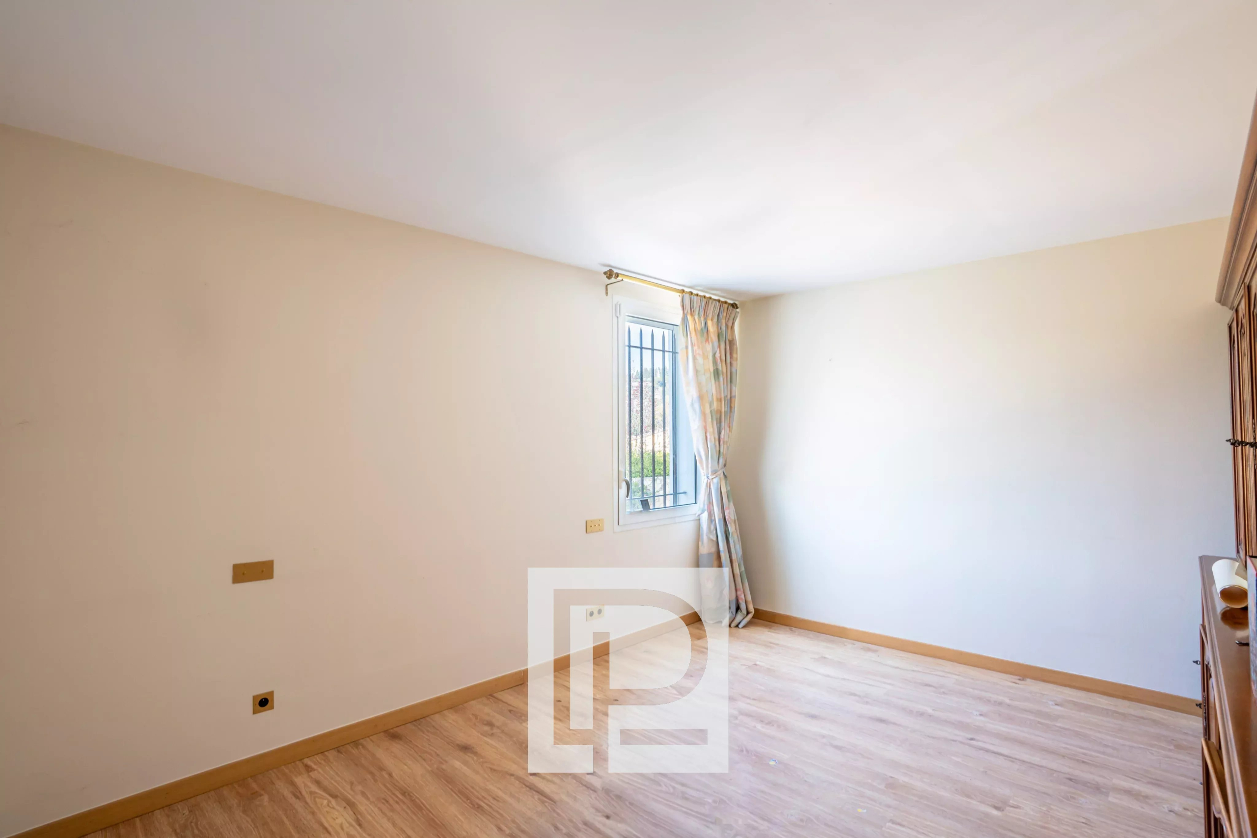 Empty beige-walled room with a light wood floor, a window with bars, and a patterned curtain pulled to the side on the far wall, plus a wooden dresser on the right.