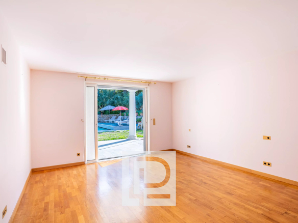 Bright empty living room with light pink walls and wooden floor, sliding glass door open to a patio with a pool and red umbrella.