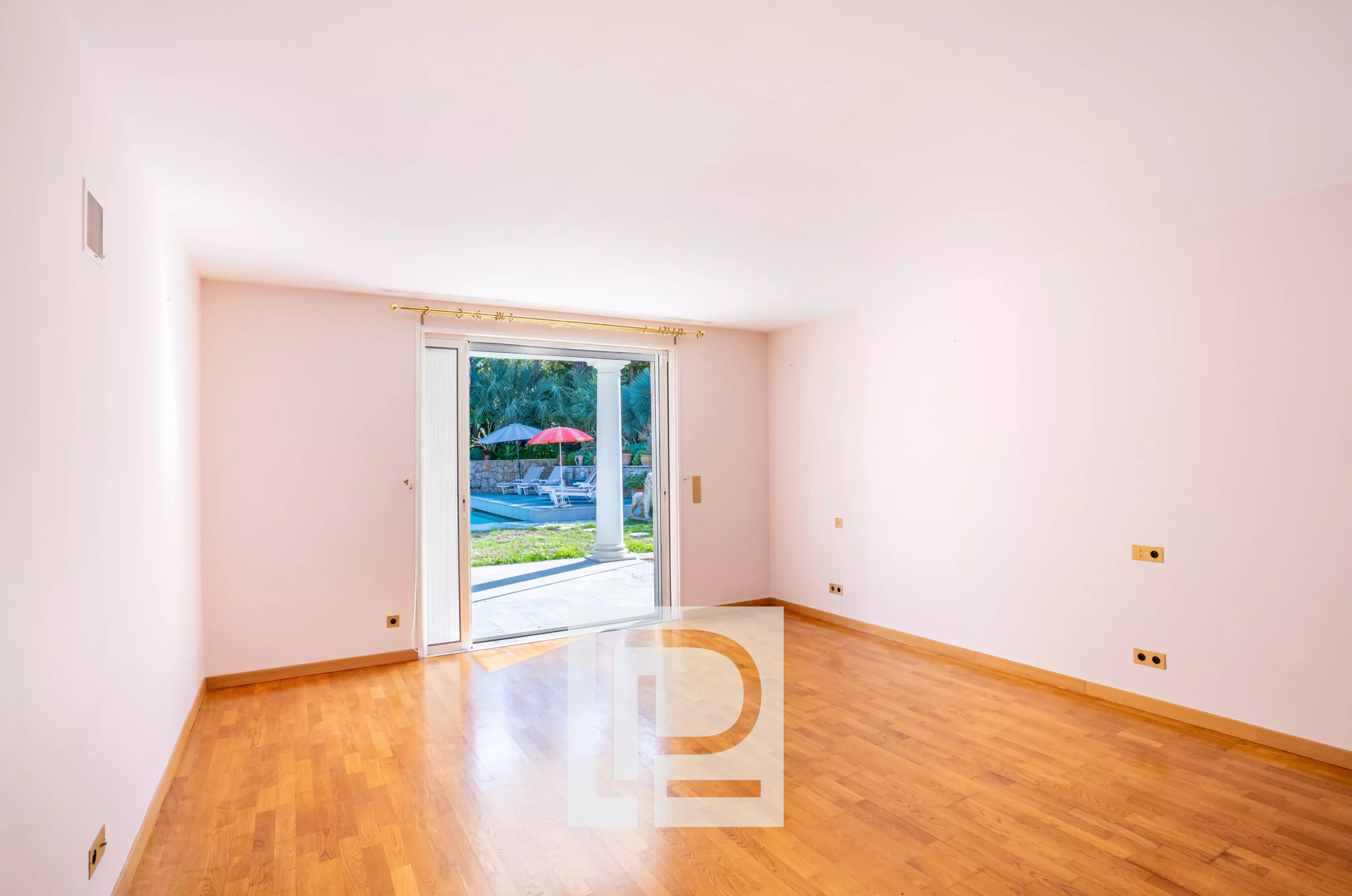 Bright empty living room with light pink walls and wooden floor, sliding glass door open to a patio with a pool and red umbrella.