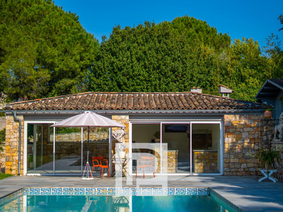 Stone house with a tiled roof and glass sliding doors overlooking a rectangular pool, a pink patio umbrella and orange chairs on a stone patio, surrounded by lush green trees under a bright blue sky