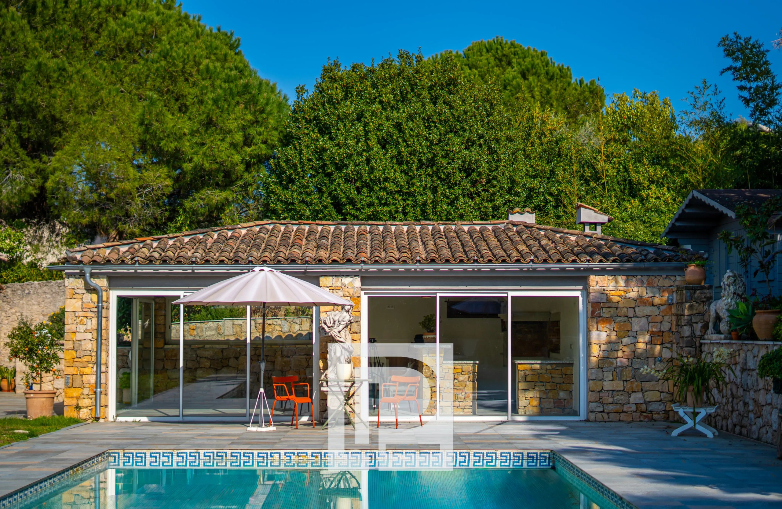 Stone house with a tiled roof and glass sliding doors overlooking a rectangular pool, a pink patio umbrella and orange chairs on a stone patio, surrounded by lush green trees under a bright blue sky