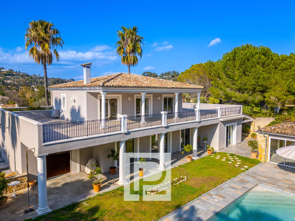 Two-story villa with white columns, tiled roof, wraparound balcony, and a rectangular pool nearby on a sunny day.