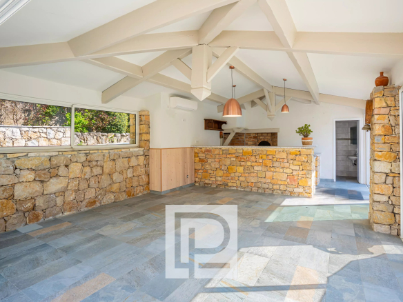 Open-plan room with stone walls, white vaulted beams, and a stone bar counter with copper pendant lights; large windows reveal a garden.