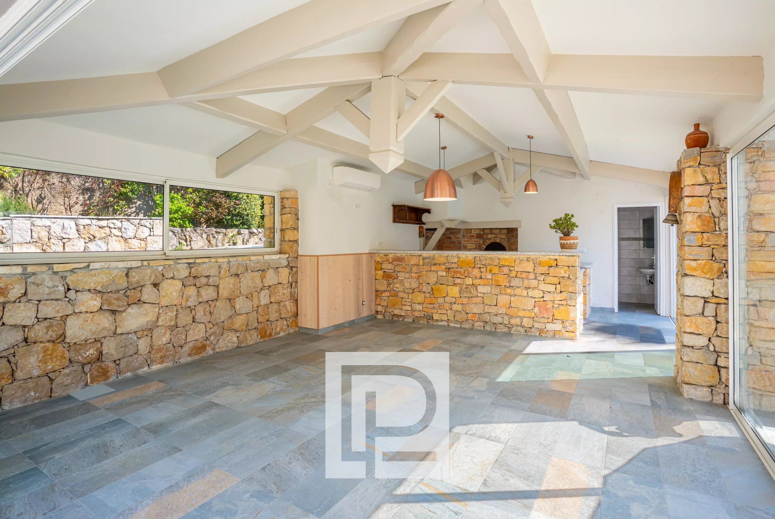 Open-plan room with stone walls, white vaulted beams, and a stone bar counter with copper pendant lights; large windows reveal a garden.