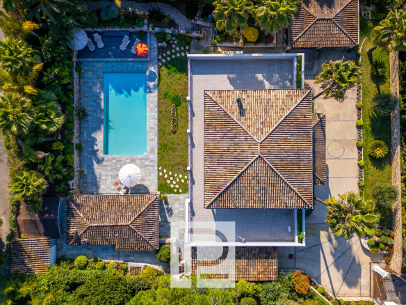 Aerial view of a villa with a rectangular swimming pool, tiled patio, and surrounding palm trees and lawn areas.