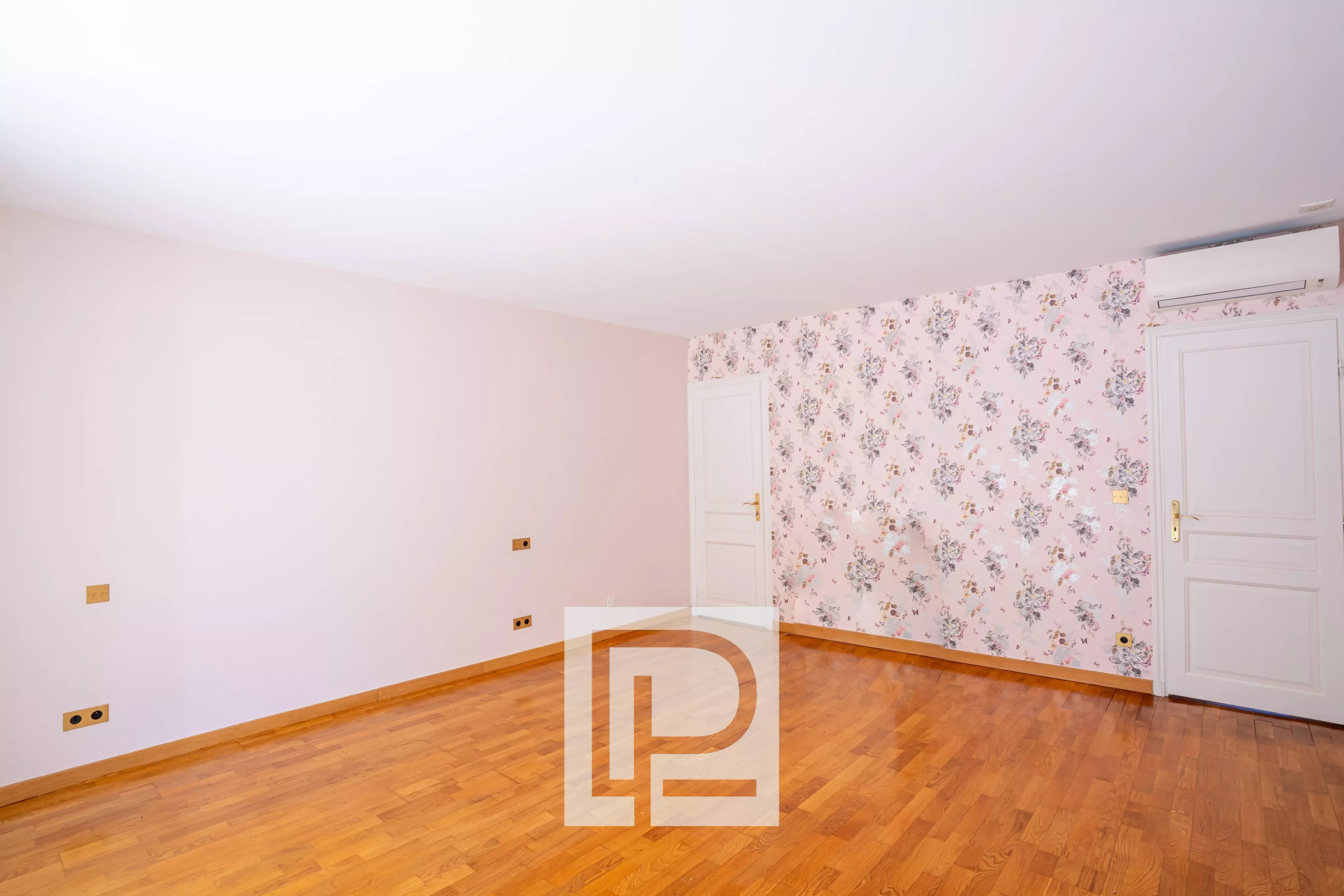 Empty room with pale pink walls, a floral wallpaper accent wall, hardwood floor, two white doors, and an air conditioner above the right door.