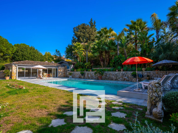 Backyard scene with a rectangular pool, stone house, and lush tropical plants under a bright blue sky.