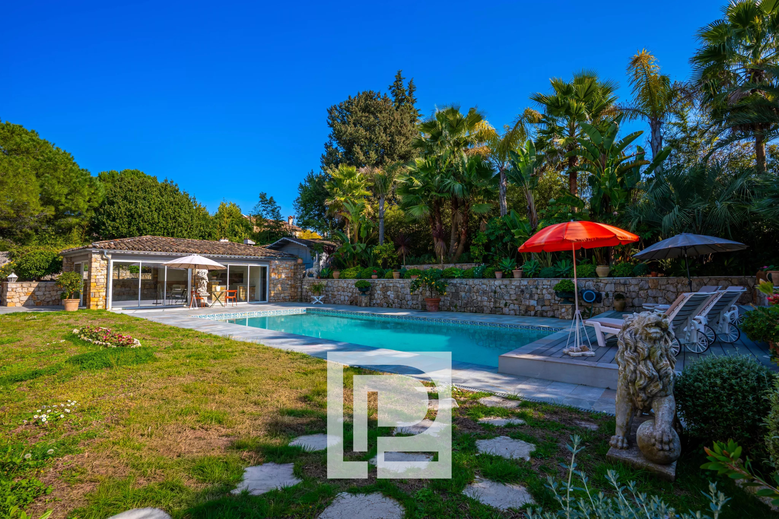 Backyard scene with a rectangular pool, stone house, and lush tropical plants under a bright blue sky.