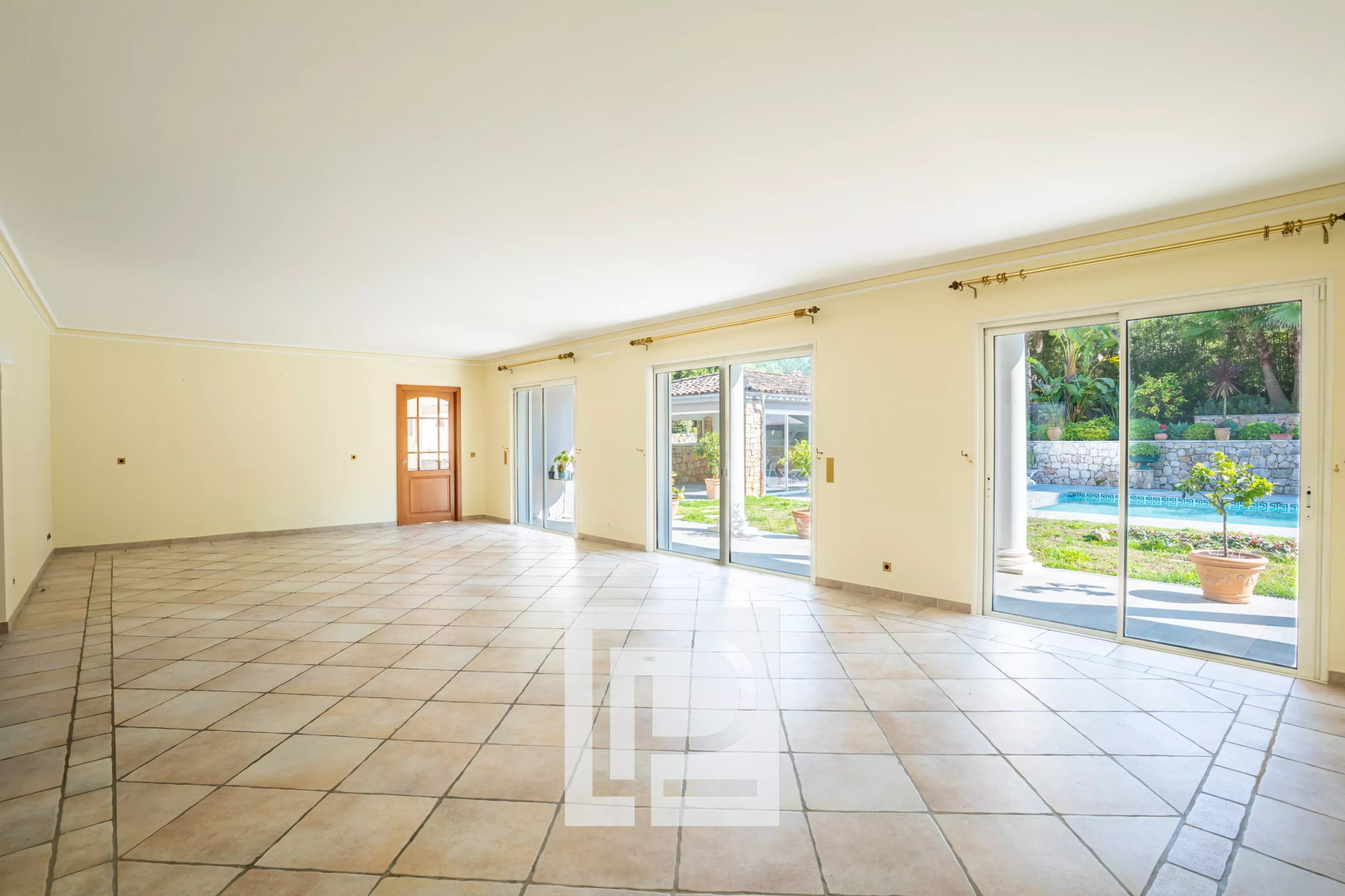 Bright empty living room with beige walls, tiled floor, and large sliding doors opening to a pool and patio outside.