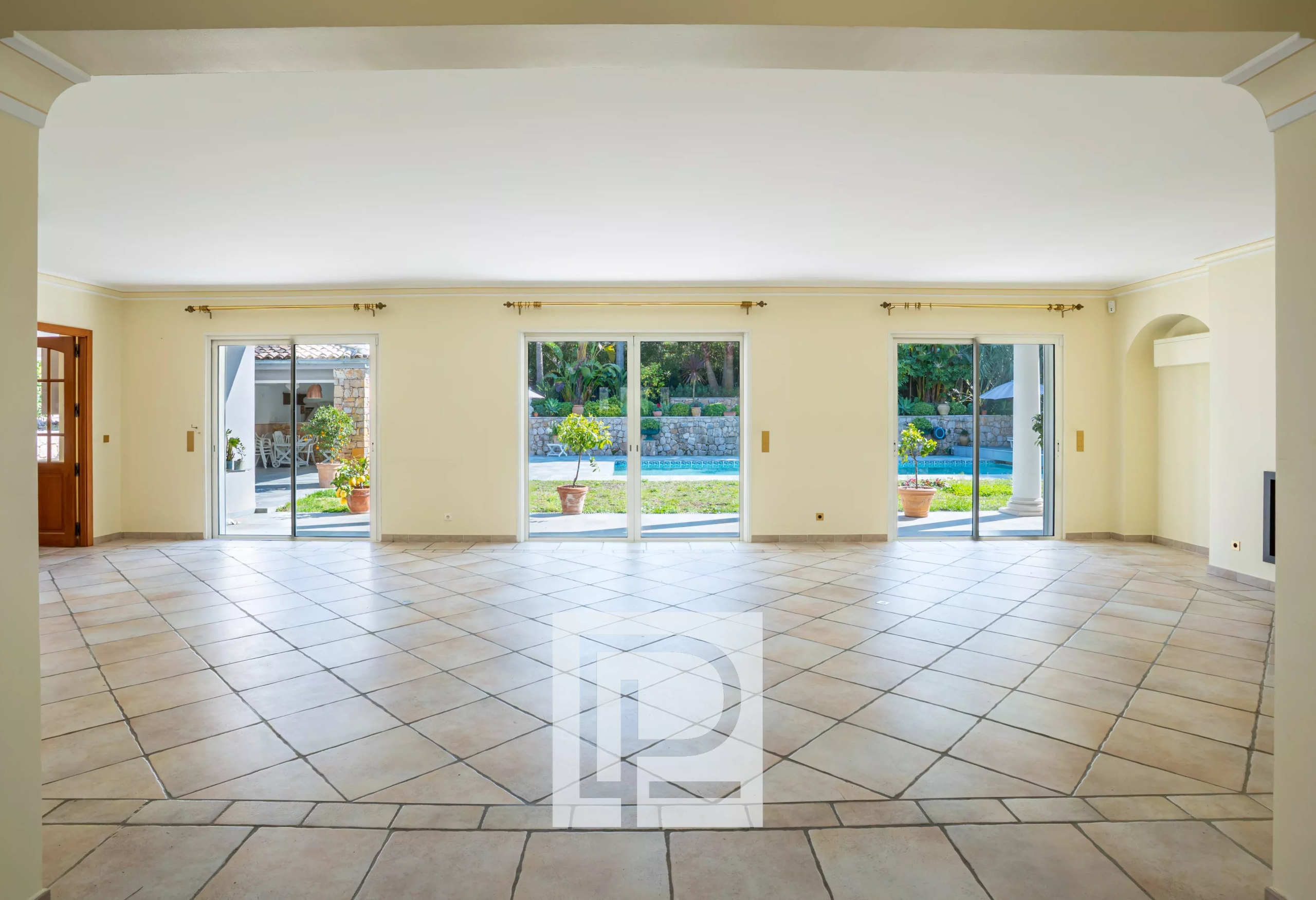Bright empty living room with tiled floor and wide sliding glass doors opening to a garden and pool area outside.